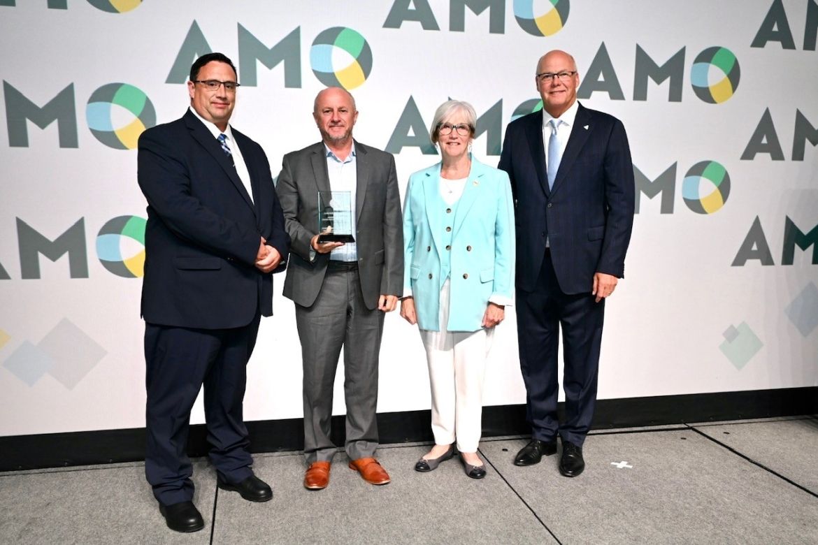 Four individuals standing against the AMO background with one individual holding an award